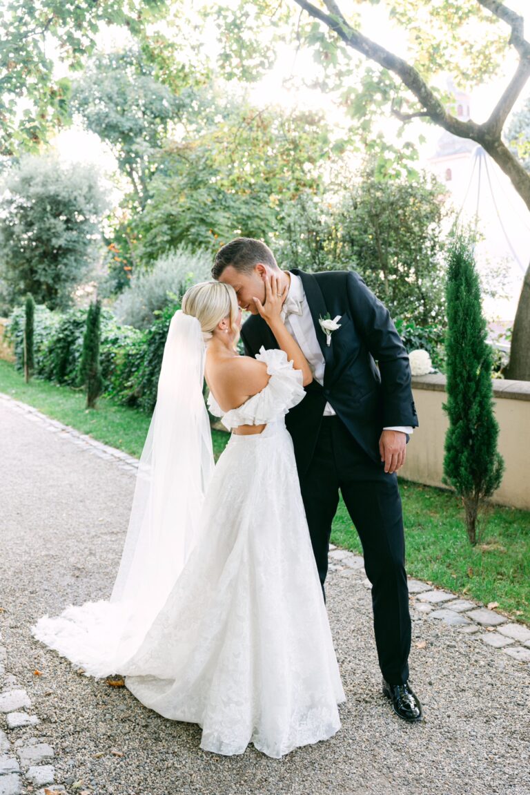 Bride and groom sharing a kiss outdoors, dressed in elegant wedding attire, surrounded by greenery.