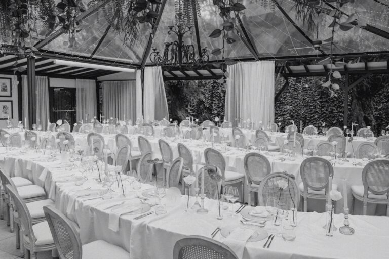 Long dining tables set with white tablecloths and elegant chairs in a glass-enclosed venue.