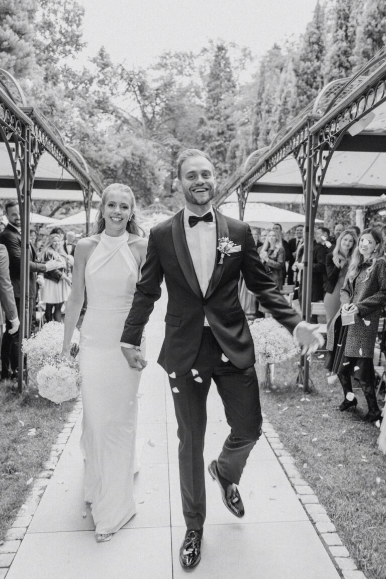 Die Braut im eleganten Kleid und der Bräutigam im Smoking gehen nach der Trauung Hand in Hand mit den Gästen im Hintergrund.