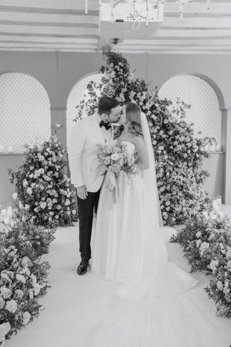 Bride and groom sharing a kiss during their wedding ceremony surrounded by floral arrangements.